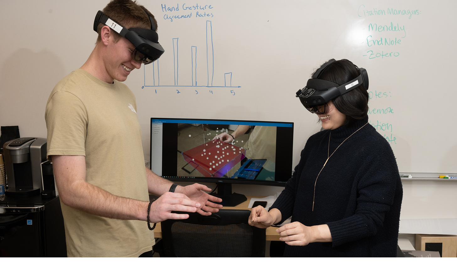 Two smiling lab members wearing augmented-reality headsets during a hand-gesture study, with a gesture agreement-rate chart drawn on the whiteboard behind them.