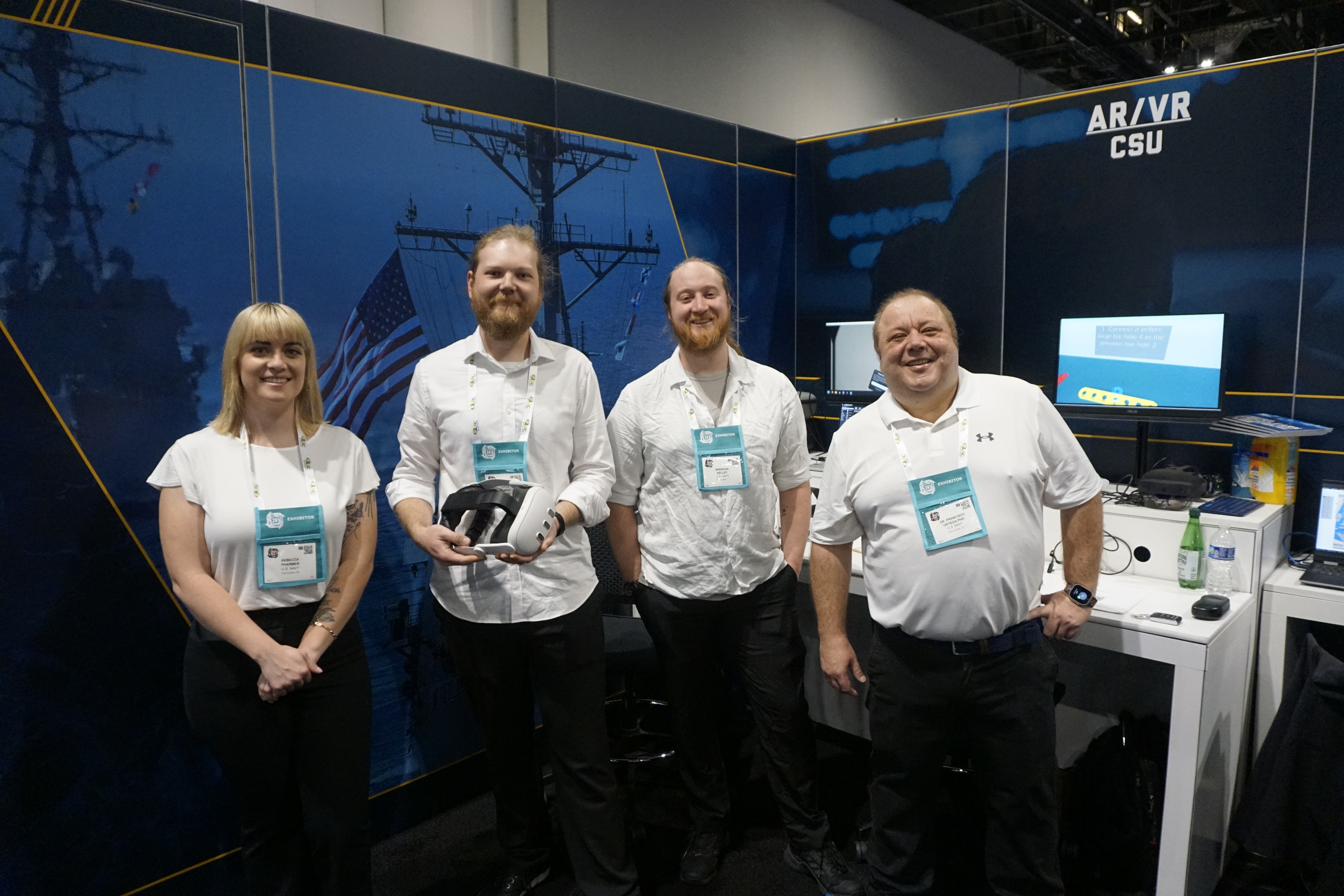 Four NUILab members at the AR/VR CSU exhibition booth, one holding a white VR headset, with naval-training demos on screens behind them.