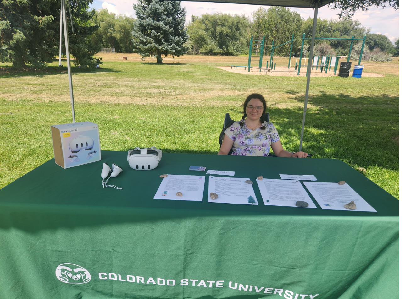 A lab member at an outdoor Colorado State University table in a park, with a VR headset and consent forms laid out for a community study.
