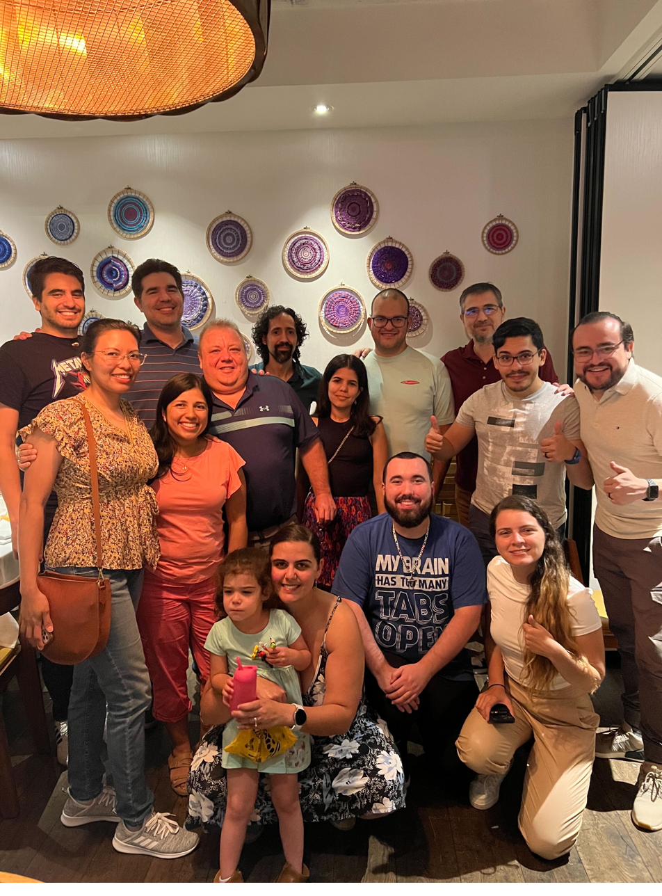 Fifteen lab members and friends posing together at a restaurant beneath a wall of colorful woven decorations.